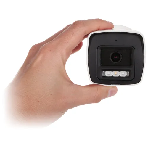 Na zdjęciu widać rękę trzymającą białą kamerę monitoringu z widocznym obiektywem i diodami LED. Kamera ma prostokątny kształt z zaokrąglonymi krawędziami.