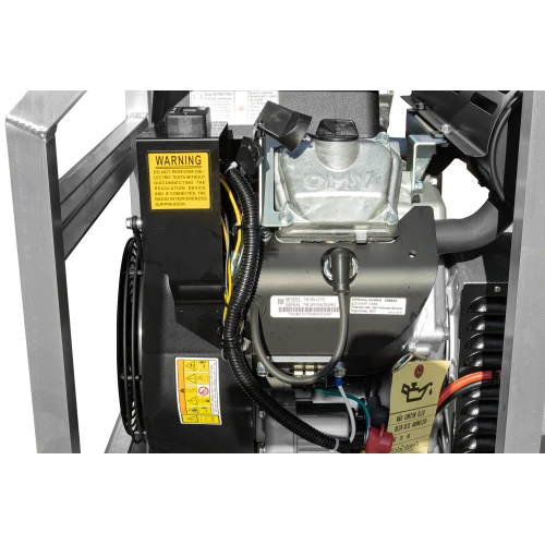 Zdjęcie przedstawia zbliżenie na silnik agregatu prądotwórczego z widocznymi etykietami ostrzegawczymi i informacyjnymi. Widać szczegóły konstrukcji, takie jak przewody i metalowe elementy obudowy.