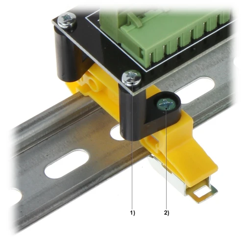 Zdjęcie przedstawia moduł elektroniczny zamontowany na metalowej szynie DIN za pomocą żółtego uchwytu. Widoczne są szczegóły montażu, w tym śruby i złącza modułu.
