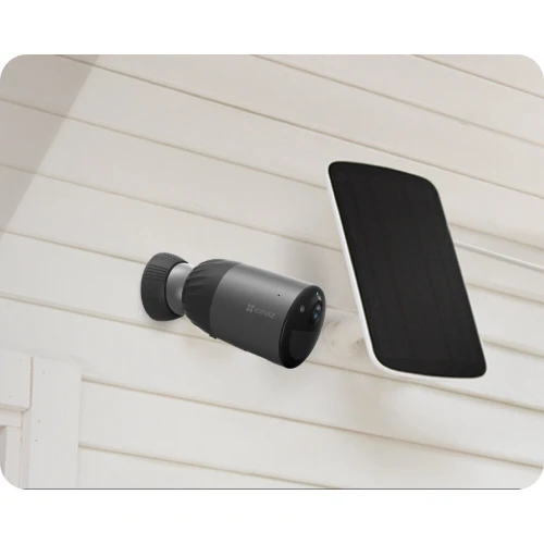 Kamera na baterie WiFi 4MPx w kolorze czarnym zamontowana na zewnętrznej ścianie budynku. Obok kamery znajduje się panel słoneczny do ładowania.