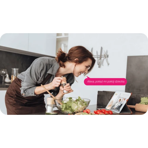 Kobieta w kuchni przyprawia sałatkę, patrząc na tablet z włączonym wideo. Na ekranie tabletu widoczny jest obraz z kamery, a obok znajduje się dymek z tekstem "Alexa, pokaż mi pokój dziecka".